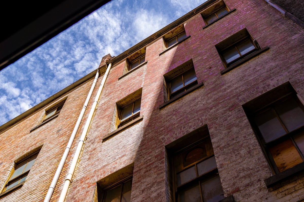 An aged brick building wall with windows and drainpipes is captured from a low angle, revealing a cloudy blue sky above.